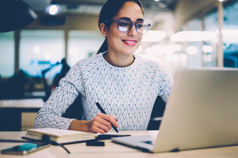 Woman sitting at desk looking and smiling at a laptop and taking notes