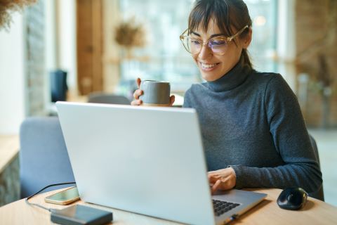 Woman sitting at a desk smiling at her laptop and holding a mug